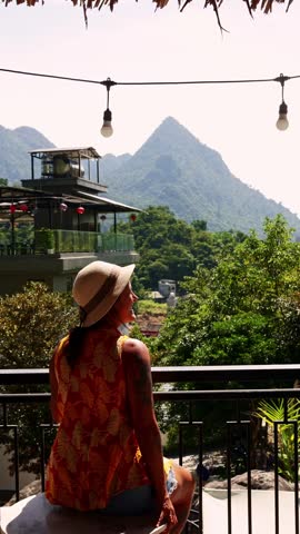 Female tourist wearing a sun hat sitting on a hotel balcony in a tropical destination, relaxing and admiring the lush green mountains and scenic landscape during her summer vacation