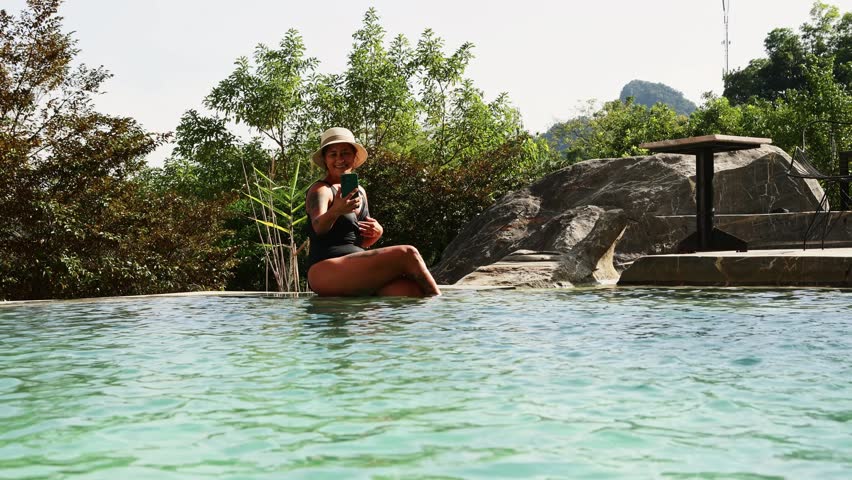 Cheerful young woman wearing a hat taking a selfie with her phone while sitting on the edge of a beautiful infinity pool at a luxury resort surrounded by tropical nature