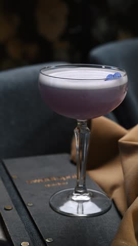 Side view of purple cocktail with foam in coupe glass. A lavender gin sour cocktail with thick white foam and a blue flower garnish in a crystal glass.
