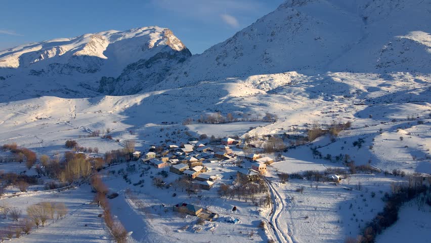 Snowy village nestled in mountain valleys. Sunlight glints off snow-capped peaks. Paths wind through quiet, frozen fields. Village homes glow warmly under winter sky