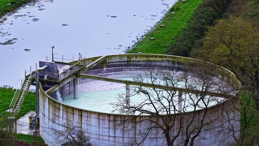 Aerial shot of a large circular industrial wastewater settling tank with metal walkways and stairs beside a muddy river surrounded by green grass and bare autumn trees on an overcast day