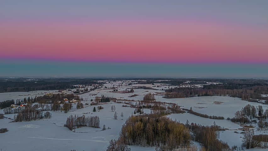 Cinematic drone shot moving forward toward a pink and blue horizon, revealing a snowy woodland and rural fields during a quiet winter dusk.