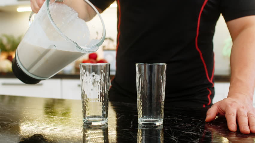 Man pours protein smoothie from blender into glass on kitchen counter for healthy nutrition. Shake with banana milk cottage cheese and whey protein supports muscle recovery weight control fitness gym.