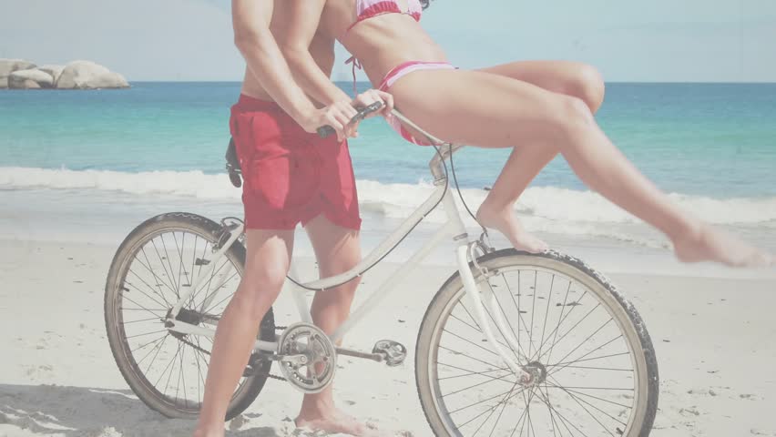 Woman climbing onto bike, man steadying, both playing, social media overlay rising around couple. Bicycle, beach, duo, romance, outdoor, advertising, lifestyle