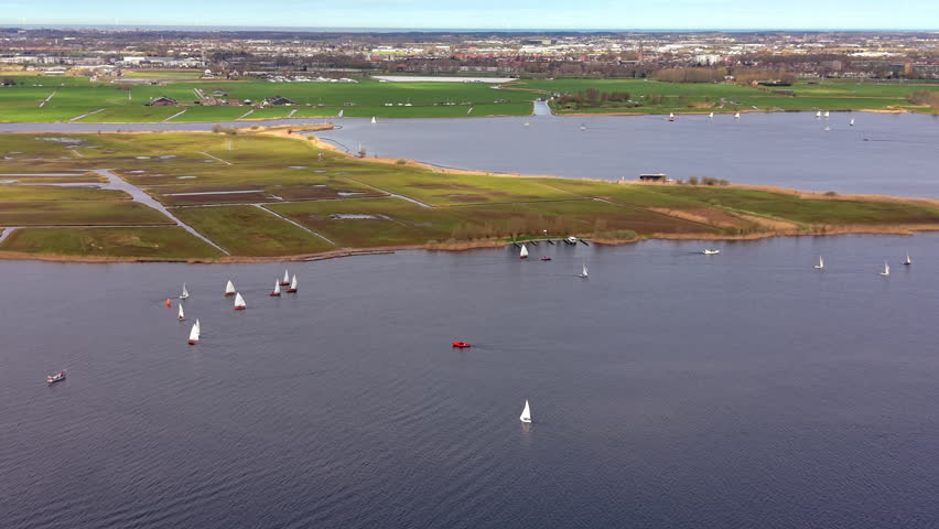 Aerial panorama of white sailboats on a blue lake near a lush green coastline with a distant city skyline. Scenic Dutch landscape featuring water sports and rural beauty under a bright sky.