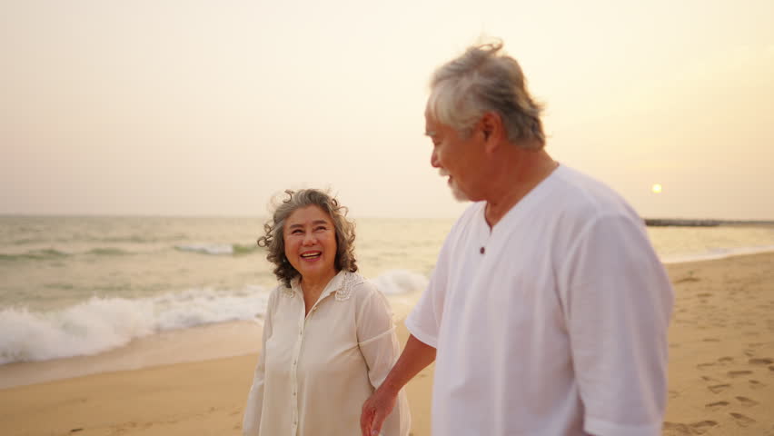 Asian senior couple holding hands and walking together on the beach at sunset, elderly enjoying romantic moment, happy retirement family lifestyle, old age love and relationship, travel nature outdoor activity by the ocean.