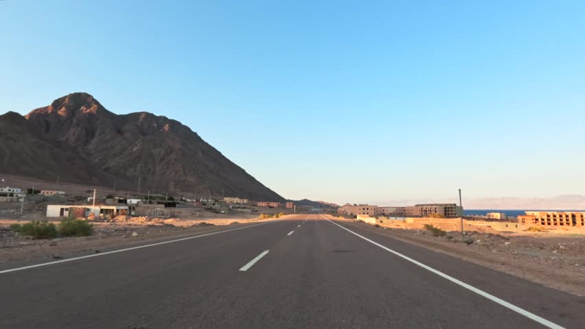 Car driving on desert highway near mountain and suburban buildings at sunset