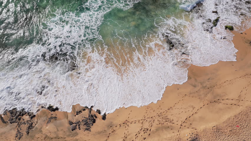 Aerial top down view of a beach with waves crashing onto the shore. Green water and white foam indicating turbulent waves. Warm color sand with footprints