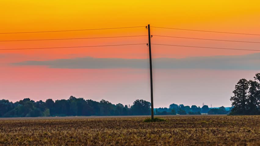 Sunrise timelapse over a rural farm field with a utility pole and power lines silhouetted against an orange dawn sky and distant treeline.
