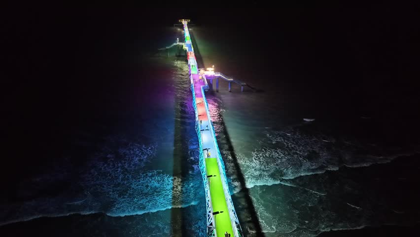 Aerial view of ocean pier at night with vibrant illumination creating stunning reflections on water, gentle waves breaking on shore, providing calm and magical atmosphere.