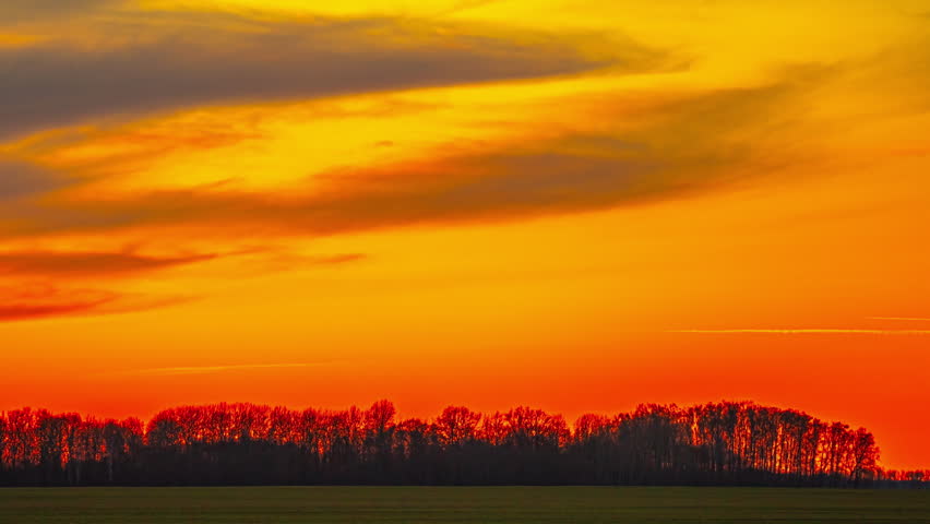 Dramatic telephoto time-lapse captures blazing diagonal cloud bands racing across fiery red and orange sky above dark silhouetted treeline and flat field in Latvian countryside