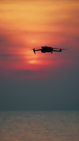 Drone silhouette hovering over sea at sunset with soft red orange sky.