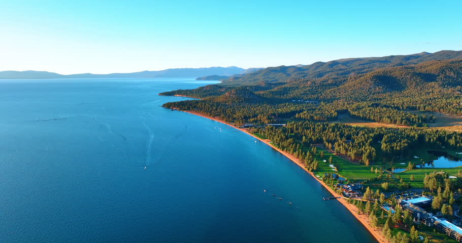 Flight over the blue vast waterscape of Lake Tahoe, California, USA. Beautiful waterfront is covered with evergreen pine tree woods.
