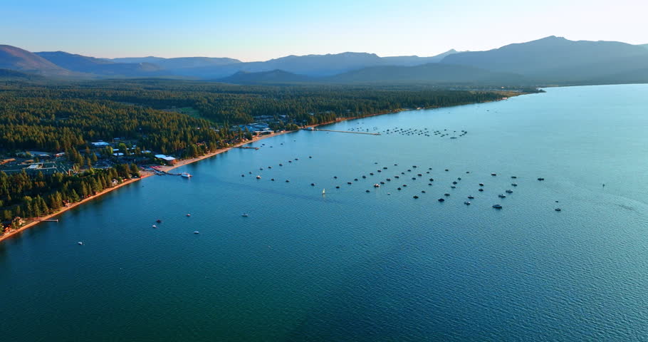 Multiple boats stand on the surface of a vast waterscape. Vast green pine tree forests cover the waterfront. Aerial view. Lake Tahoe, California, USA.
