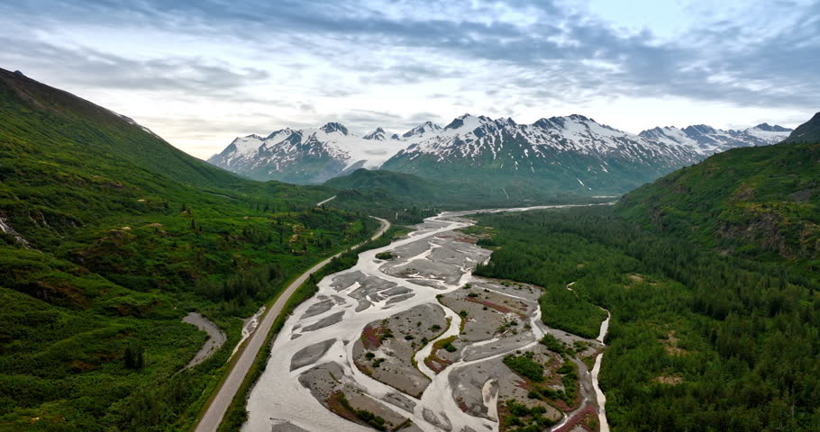 Shallow branching river and parallel highway cross the vast valley. Aerial perspective on the spectacular green mountains and snow-capped rocks. Alaska, USA.
