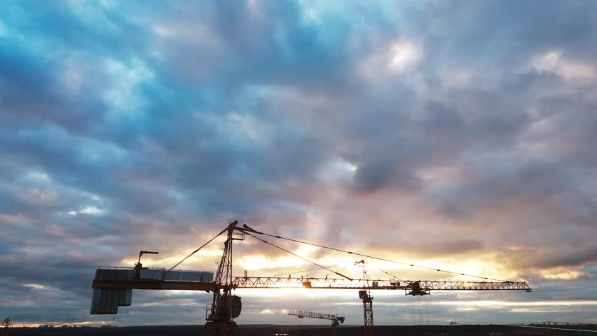 Dramatic clouds glow behind industrial cranes. Silhouetted structures stand against a vibrant sky. Sunlight breaks through dense storm clouds. Construction site bathed in twilight