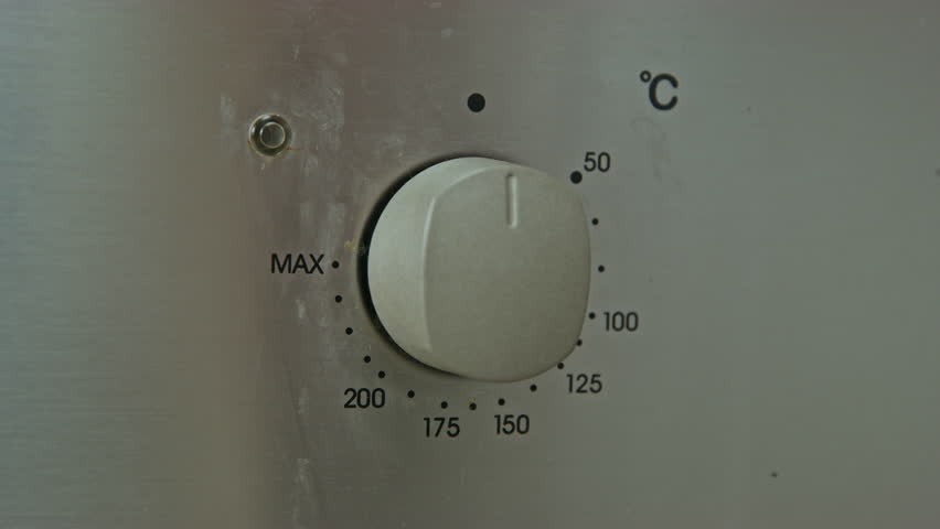 A human hand turns the oven temperature control dial on a stainless steel panel to the MAX mark, demonstrating detailed settings for precise control of the cooking process.
