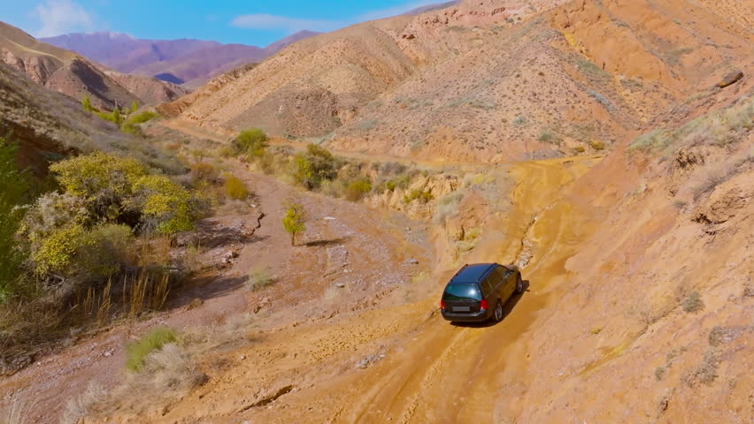 Dark colored car navigates winding dirt road in rugged mountainous terrain. Dry vegetation and canyons create an adventurous atmosphere under daylight. Following drone view.