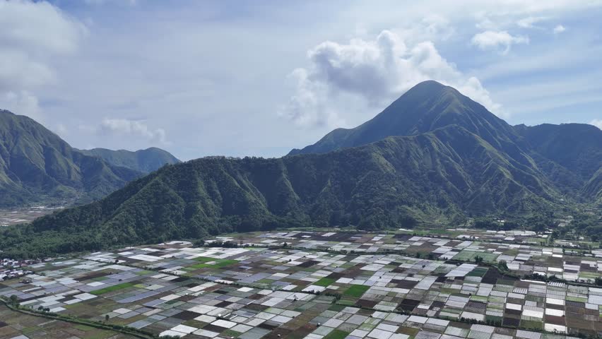 Scenic aerial view of vast agricultural fields nestled between lush green mountains under a bright sky with clouds, showcasing rural farming and natural landscape.