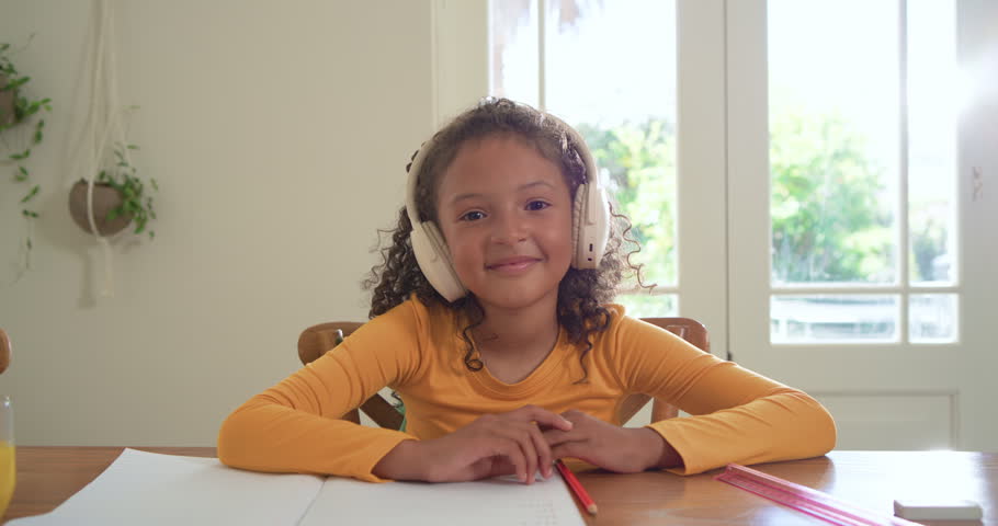School-aged girl sitting at wooden table, smiling and waving during remote lesson with headphones. Student, study, home, daylight, windows, desk, education