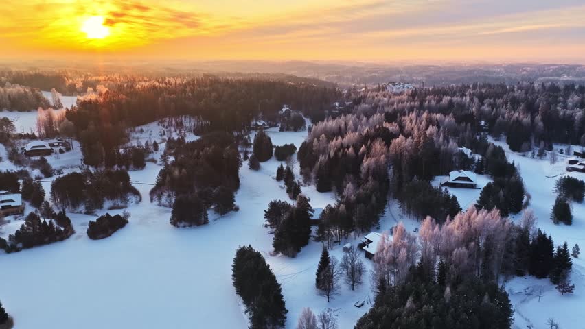 Aerial winter sunset over snowy countryside with dark pine forest, scattered cabins and frosty trees, glowing golden light across a quiet rural landscape.