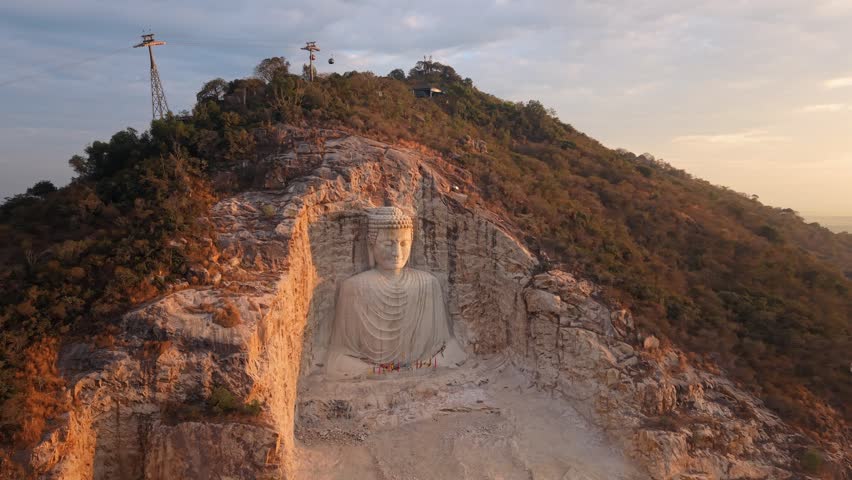 Drone orbiting shot of a large seated Buddha figure carved into a rocky cliff niche, with cable cars, colored offerings at the base, sparse hillside trees, warm late afternoon light.