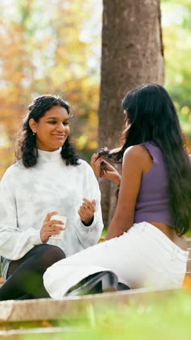 Two beautiful young indian friends having a pleasant conversation while sitting in a sunny park. They are smiling and gesturing happily
