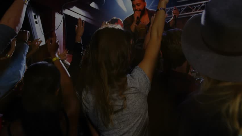 Swaying woman in gray top raising arms at club, singer singing holding mic, golden overlay. Crowd, performer, stage, microphone, venue, spotlight, smartphones