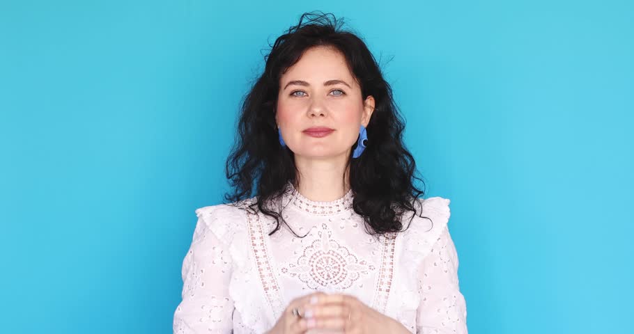 Young woman in white lace dress praying with hope, holding hands in namaste gesture with eyes closed then looking up with surprise and smiling on blue studio background