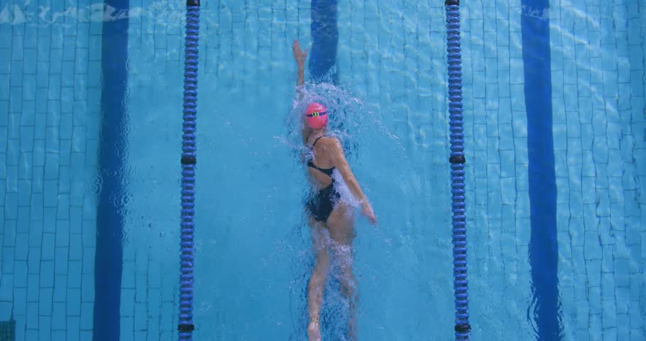 Woman swimmer starting lap, doing backstroke and kicking for sports, aligning with blue lane ropes. Athlete, aquatic, wake, splash, reflections, tiles, competition