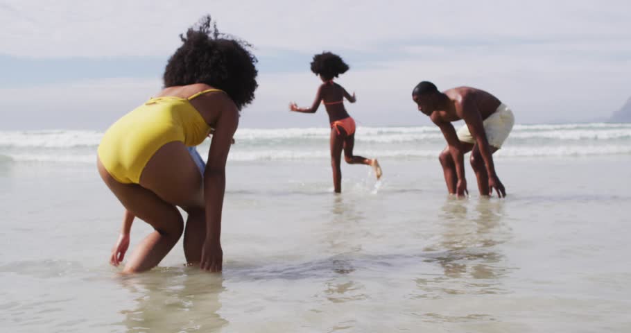 Vertical video: Running girl in orange suit to adults on shore to play, splashing with ID tags. Beach, family, ocean, waves, sand, vacation, playful