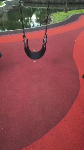 A black swing hangs from chains on a red playground surface near a pond with green grass.
