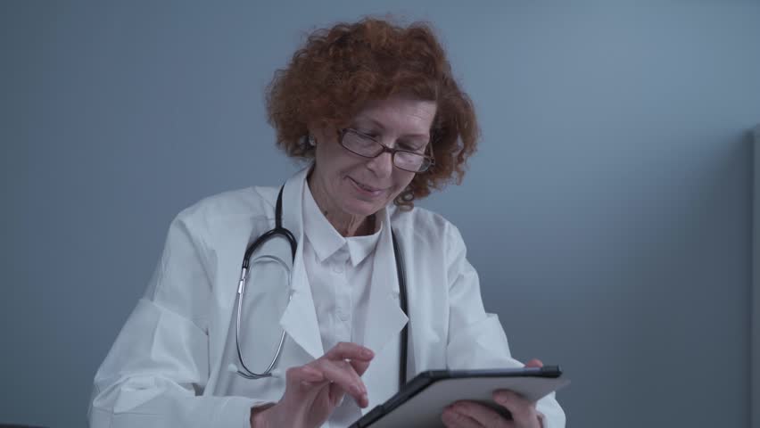 Medicine and healthcare concept. Portrait happy senior female doctor holding patient chart on digital tablet working at table in hospital office. Remote telemedicine app in hospital working online.
