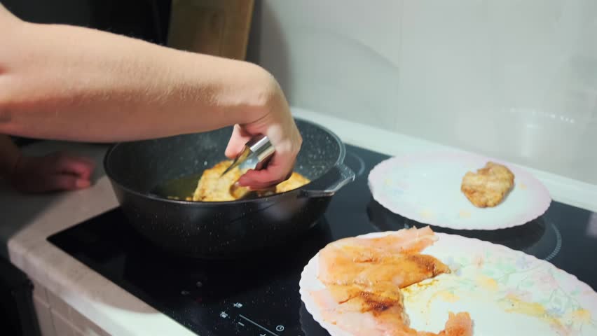 Close-up shot of a person frying chicken fillets in a non-stick pan on a modern electric stove. Homemade cooking process with focus on textures, sizzling oil, and golden crust.