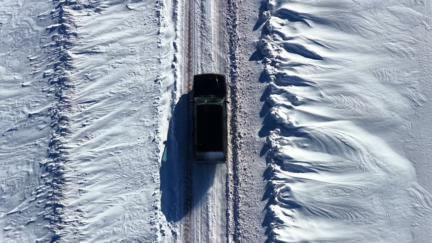 Top-down drone view of a dark car driving along a narrow snow-covered rural road between wind-shaped snowbanks in bright winter daylight.