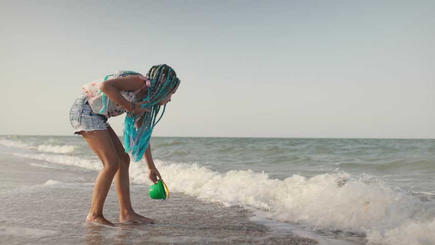 A girl with african braids in a summer costume plays on the beach with shells near the sea with waves under sunny sunset
