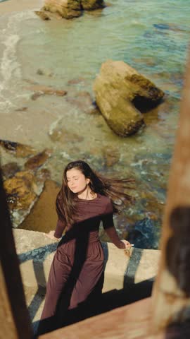 A woman sits on a stone ledge above a rocky seashore, enjoying the sun with her eyes closed. Coastal, relaxation, ocean, nature, mindfulness, peace, freedom, summer, wellness, travel, solitude