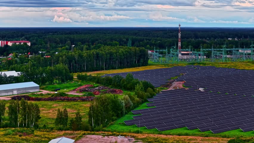 Solar panel field beside electrical substation and forest under cloudy sky, aerial