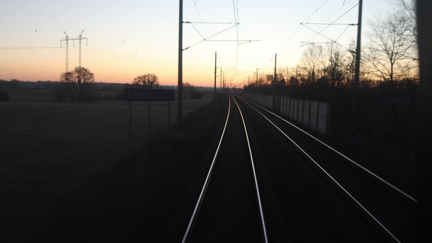 Railway tracks stretch into sunset horizon through quiet countryside with power lines silhouettes and soft evening light creating calm travel mood