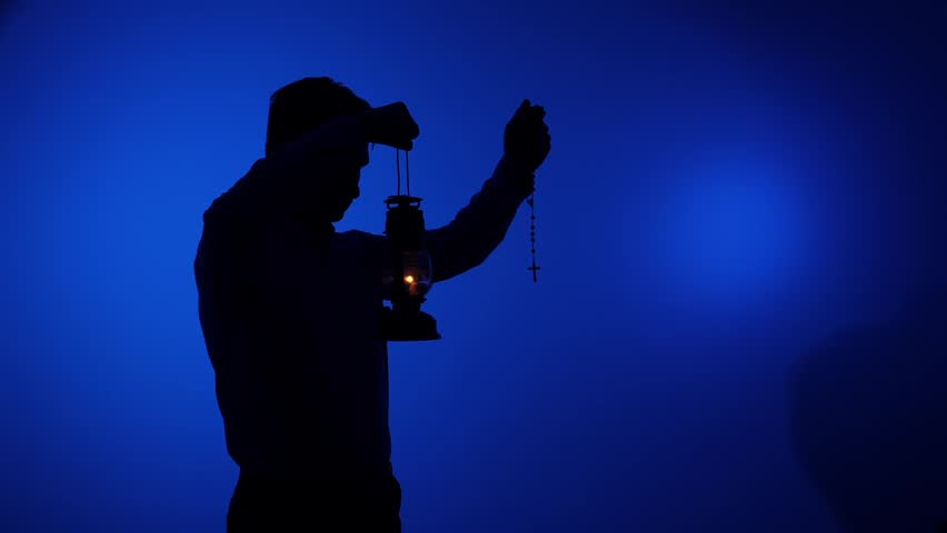 Silhouette of a person holding an old lantern illuminating the path and a rosary, symbolizing spiritual guidance, prayer, devotion, and seeking faith in times of darkness against a blue background