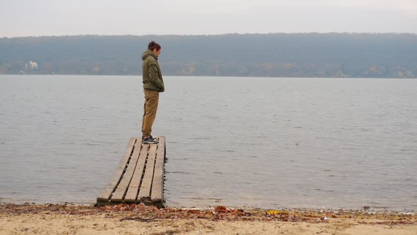 Pensive young man in a jacket standing alone on the edge of a small wooden pier, thoughtfully looking out at the calm water of a lake during a cloudy and melancholic autumn day