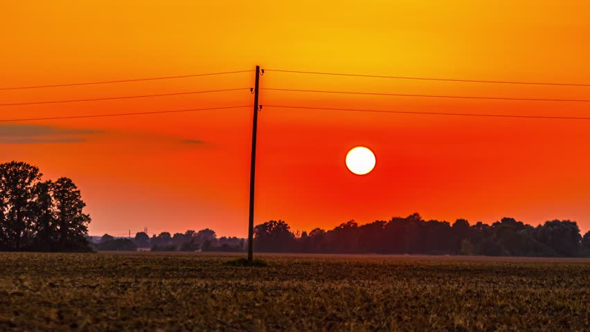 Orange sunset over rural farmland with power pole and glowing sun on horizon