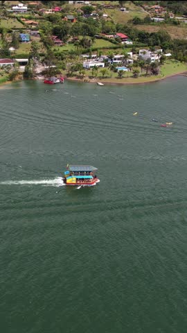 Aerial view of a double-decker party boat cruising on a vast lake near a lush, populated shore