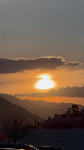 Bright sun setting behind mountains and clouds. Orange and yellow sky at dusk. Silhouette of a palm tree on the left. Dark mountain ridges in the background. Natural landscape at twilight