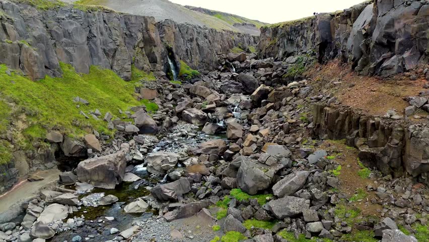 Small rock canyon along Hengifoss waterfall in Iceland with spring water flowing down rocky slope, basalt cliffs with moss and grass patches in volcanic landscape, falling rocks, showing stream water erosion, cinematic drone video