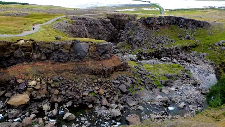 Small rock canyon along Hengifoss waterfall in Iceland with spring water flowing down rocky slope, basalt cliffs with moss and grass patches in volcanic landscape, shwoing water erosion, grasslands and lake in the background, cinematic drone video