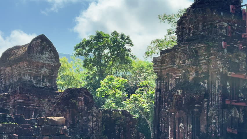 Ancient Cham Hindu temple ruins at My Son Sanctuary near Hoi An, Vietnam. Weathered red brick towers rise against lush jungle mountains and blue sky.