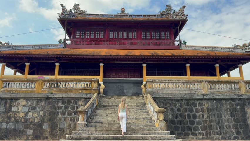 Woman walking up stone stairs toward historic imperial building in Hue, Vietnam. Traditional architecture of the Imperial City with ornate details and cultural heritage.