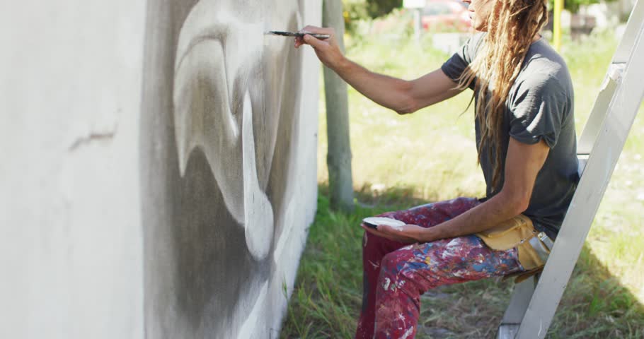 Video of caucasian male artist with dreadlocks sitting on ladder painting whale mural on wall. Freedom, creativity, inclusivity and hobbies concept digitally generated video.