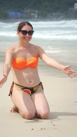 Mother Toddler Playing On Beach, Sunny Vacation Scene With Mother Kneeling In Orange Swimwear Lifting And Hugging Child In Colorful Rash Guard And Sunhat, Ocean Waves Crashing, Sandy Shore Footprints